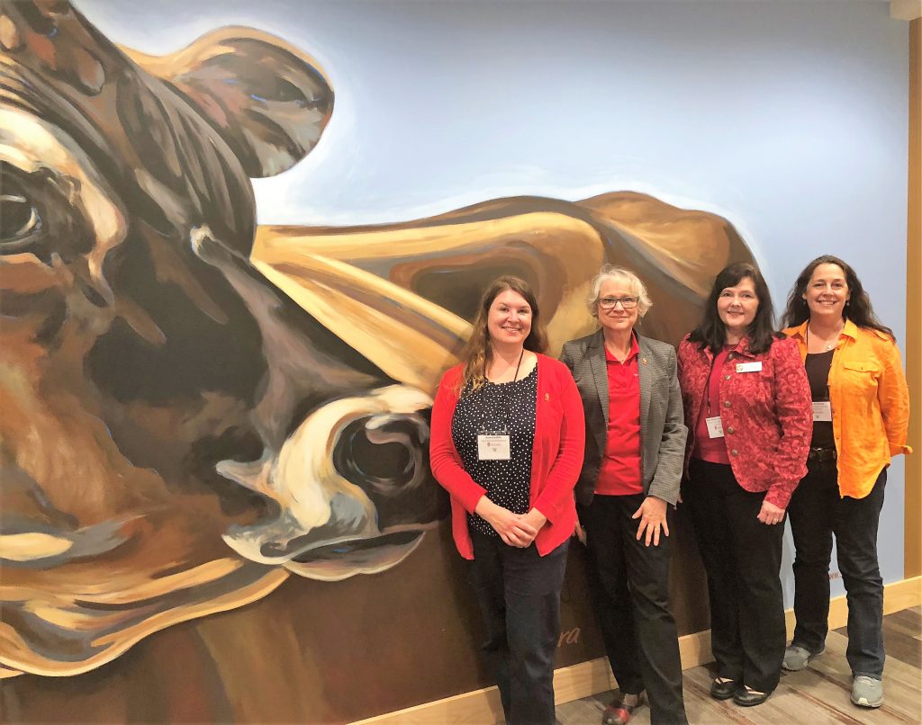Four women facing the camera smiling and posing in front of a cow mural