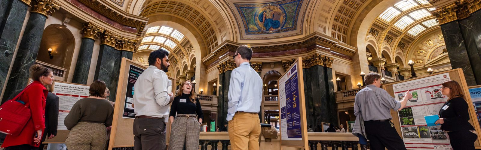 People viewing and discussing display boards in the ornately decorated rotunda of the Wisconsin State Capitol.
