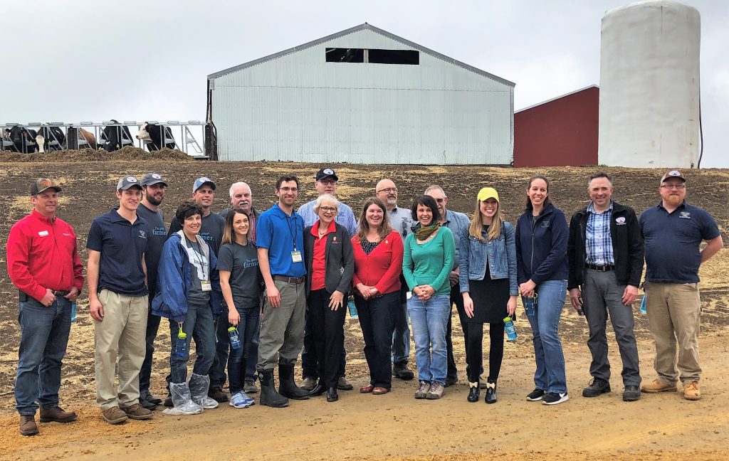 Large group photo of men and women smiling and facing the camera. Outside, near a barn