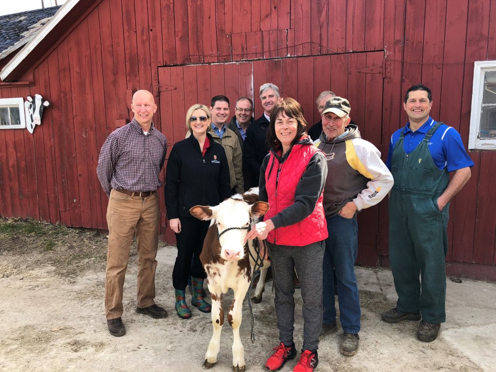 Small calf is being held by a woman and other adults posing and smiing for camera in front of a redbarn