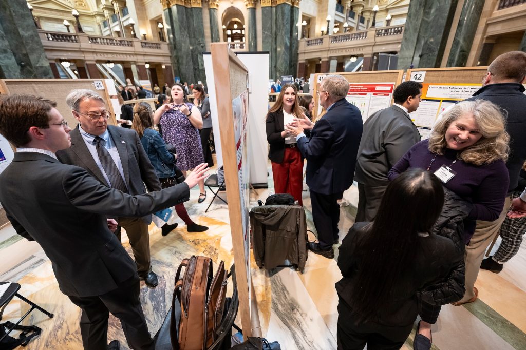 Presenters and attendees converse around display boards during a research poster session held inside a the Wisconsin State Capitol.