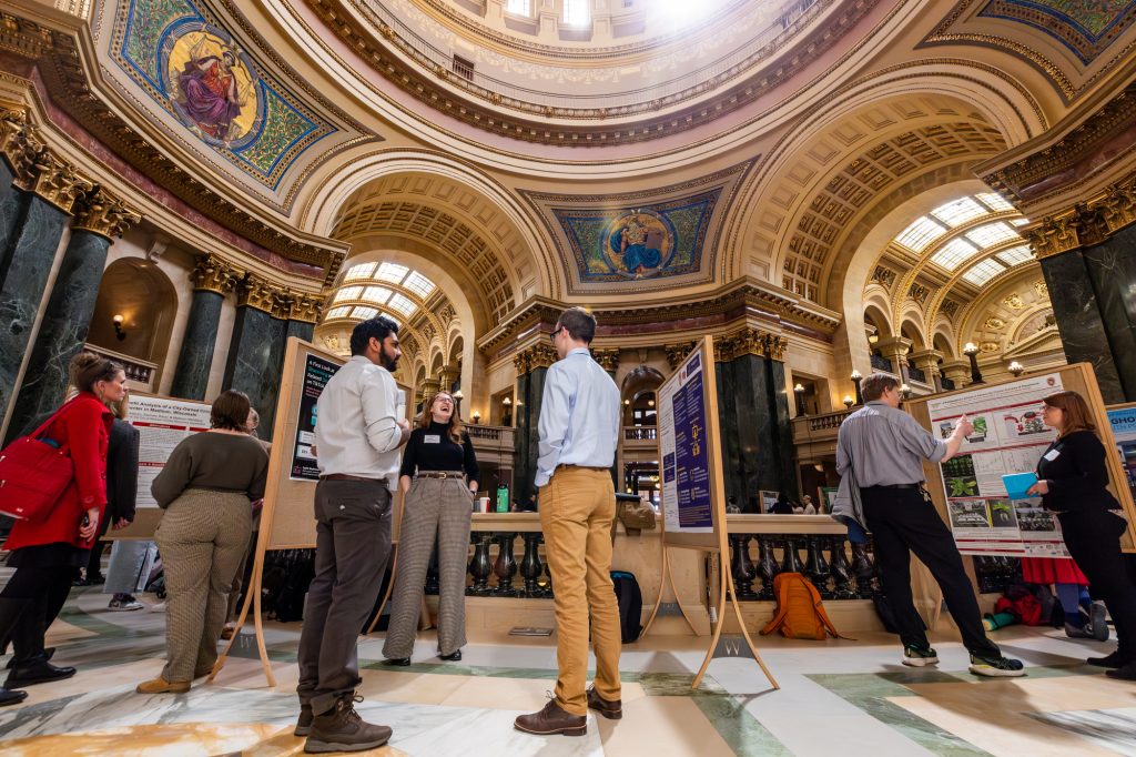 People viewing and discussing display boards in the ornately decorated rotunda of the Wisconsin State Capitol.
