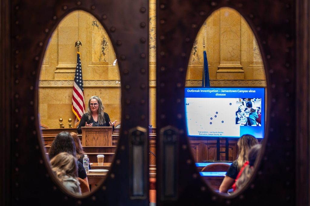 An individual presents in front of an audience in a formal chamber setting with an American flag in the background and a slideshow on the screen. The camera captures the scene through the windows of a door.