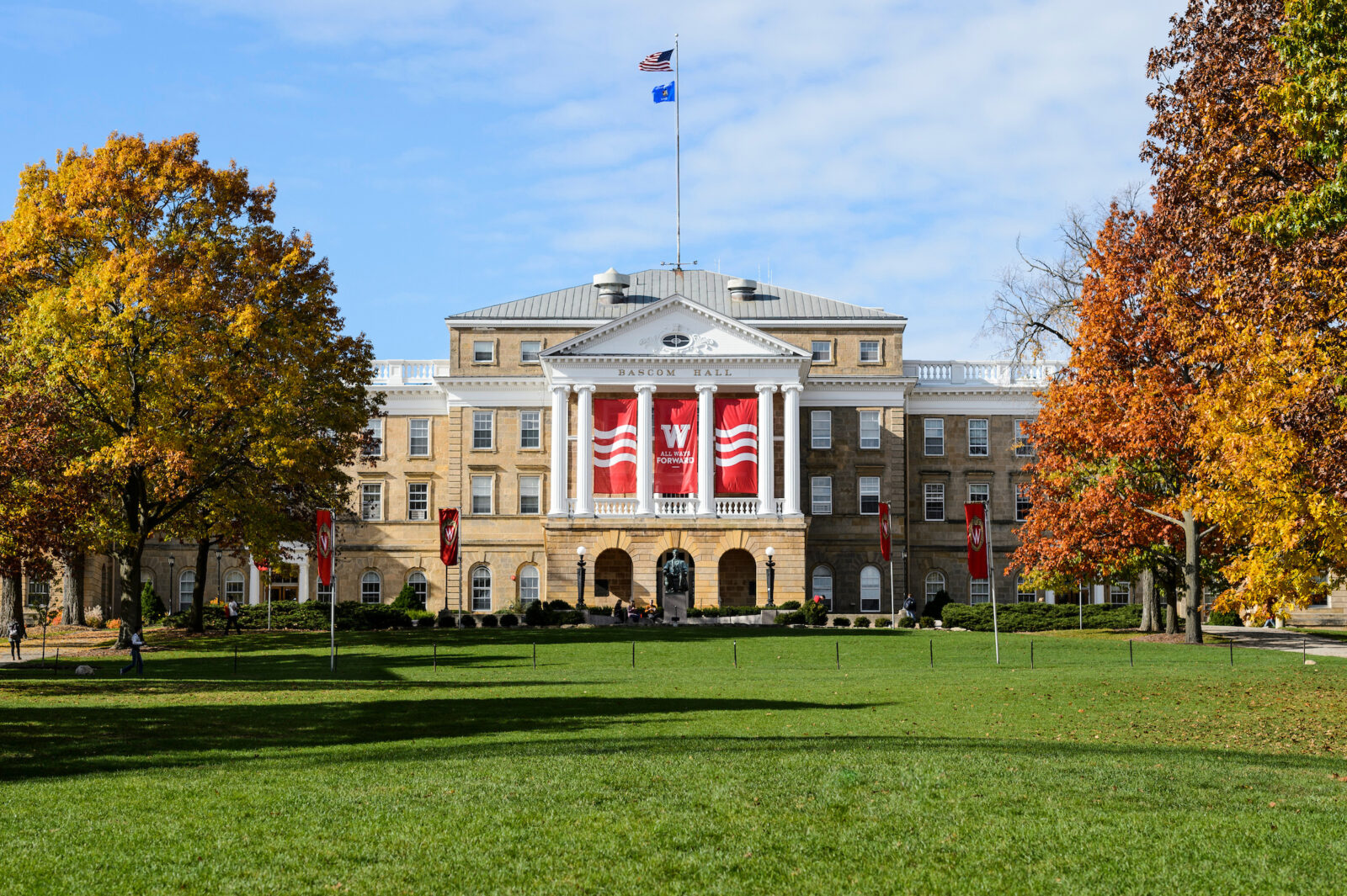 An autumn view of Bascom Hall at the University of Wisconsin-Madison, featuring a large green lawn in the foreground and trees with vibrant fall foliage framing the historic building.